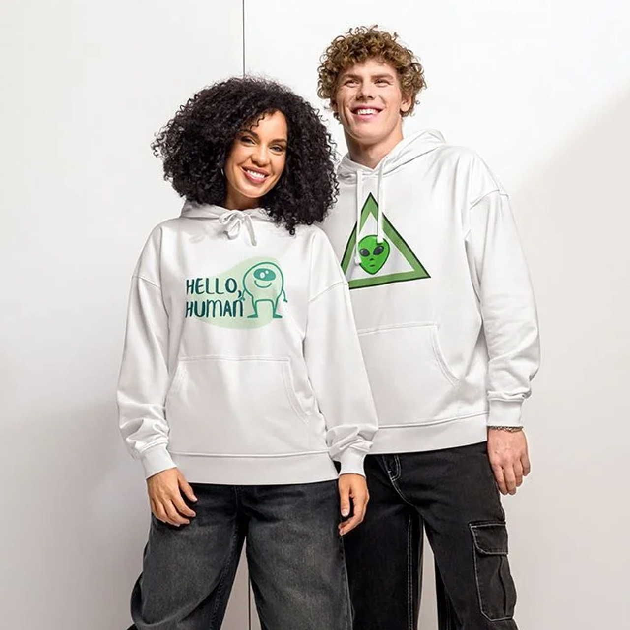 Two people smiling and wearing white hoodies. The left hoodie features a cute alien with "Hello, Human." The right hoodie has a triangle with an alien face.